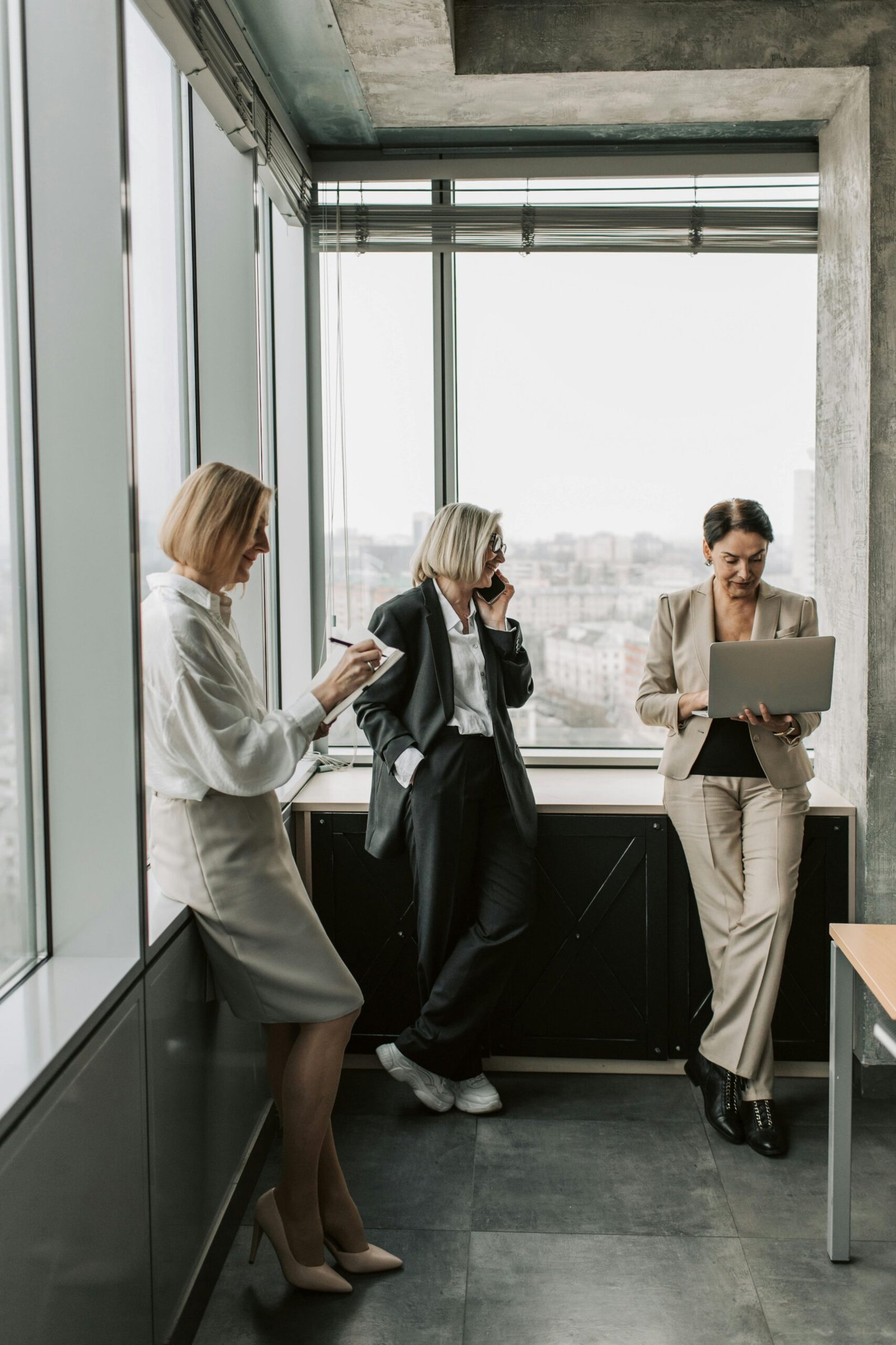 Three businesswomen in a modern office setting, working with a laptop and taking notes.